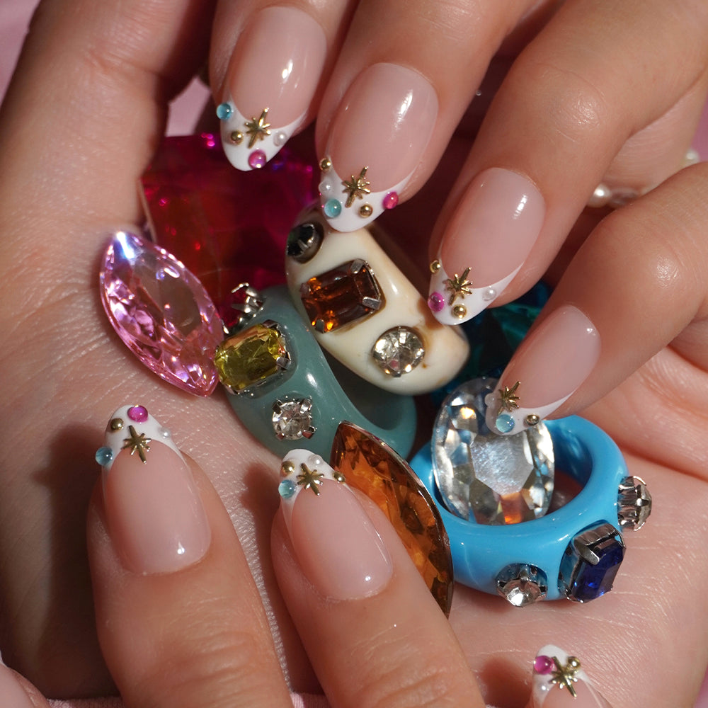 Hands with nude, almond-shaped glamnetic Moonstone press-on nails featuring white French tips, colorful gems, and gold star accents. The fingers are adorned with chunky, gem-encrusted rings and large, colorful gemstones, creating the look of a dazzling jewel-encrusted mani. - Product image