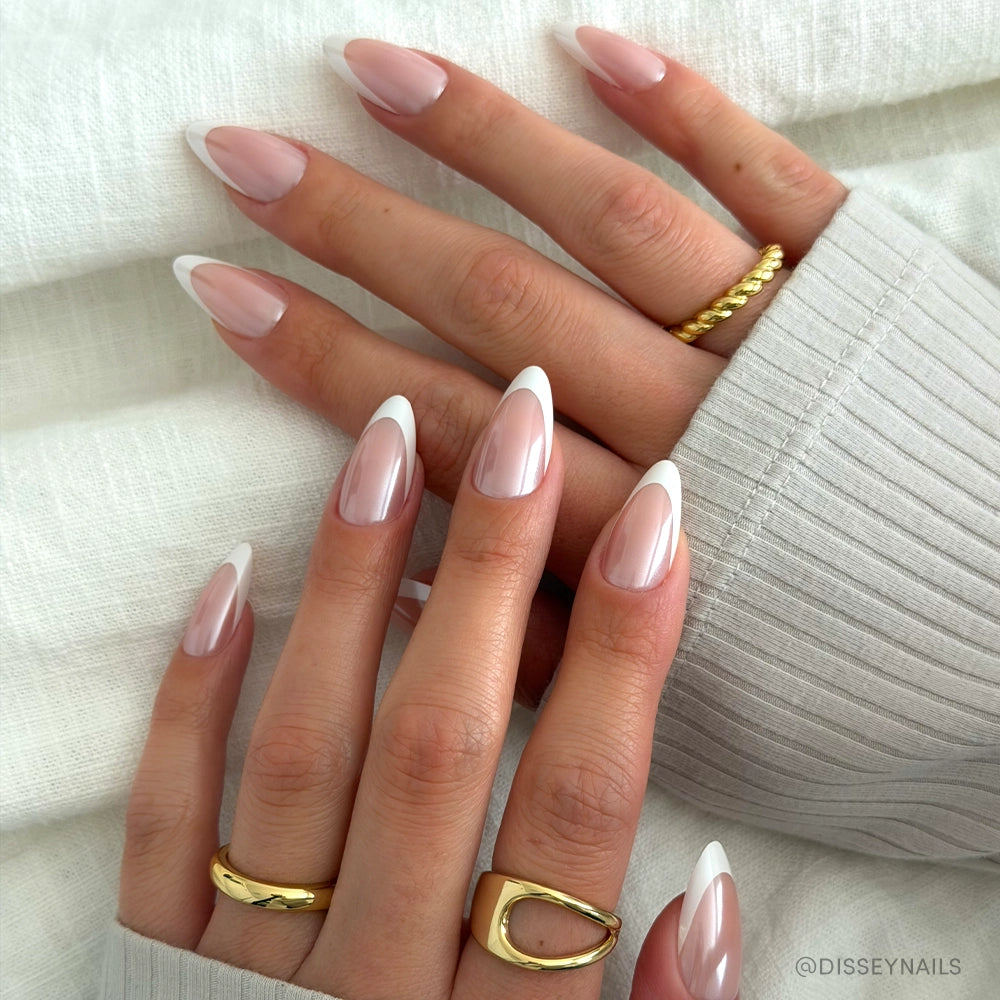 A close-up of hands with long, almond-shaped nails in glamnetic's Mother Pearl, showcasing a classic French tip and glossy finish. The hands wear gold rings and rest on textured white fabric.
