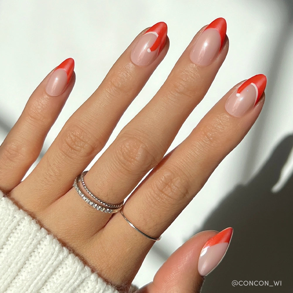 A hand with glamnetic’s Mrs. Claus glazed almond nails—nude base and red-orange abstract tips—adorned with two silver rings and a white knit sweater sleeve, creating a chic, festive look. - Product image