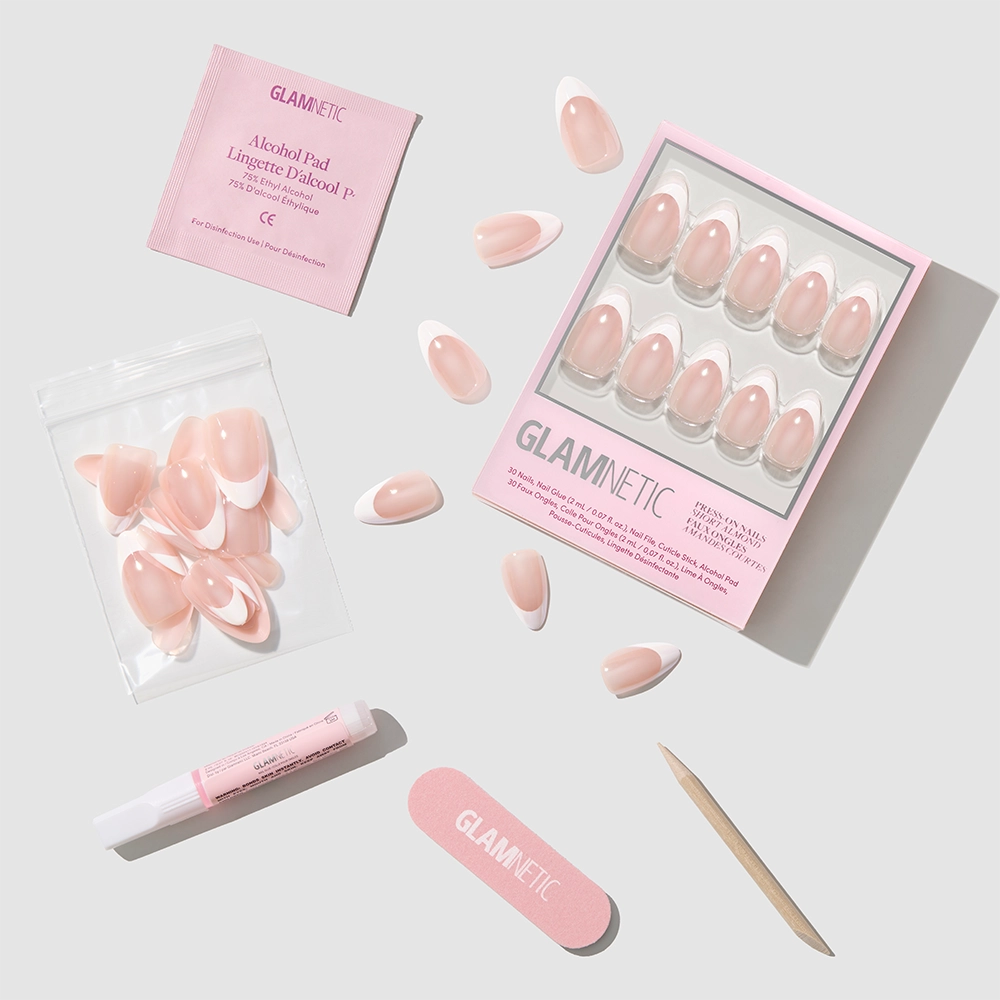 A set of Glamnetic "Mrs." pink and white French tip nails in a short almond shape come with nail glue, alcohol pad, nail file, cuticle stick, and extra nails, all neatly arranged on a light background. Packaging displays the Glamnetic logo. - Product image