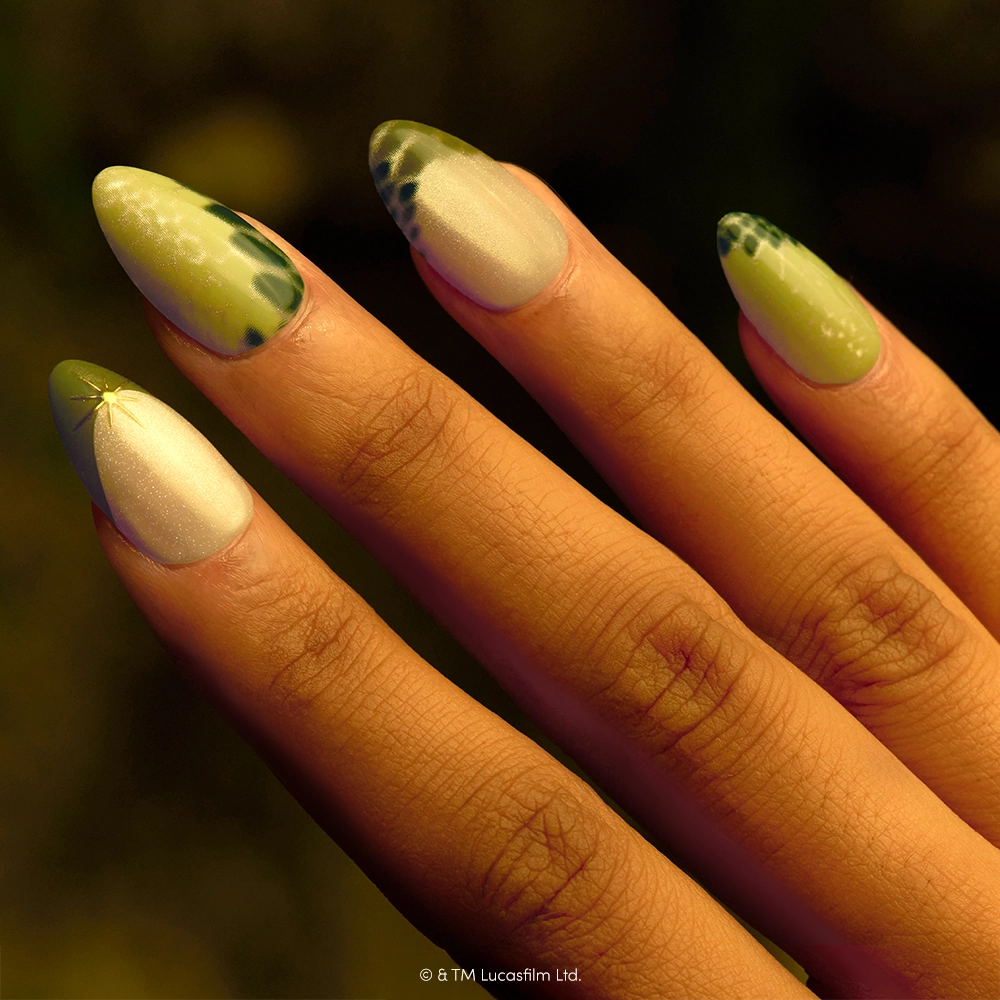 Much To Learn, You Still Have - A hand with long, almond-shaped nails painted in green and white, featuring scales and Yoda-inspired Star Wars designs, wearing the “Much To Learn, You Still Have” press-on nails by glamnetic.