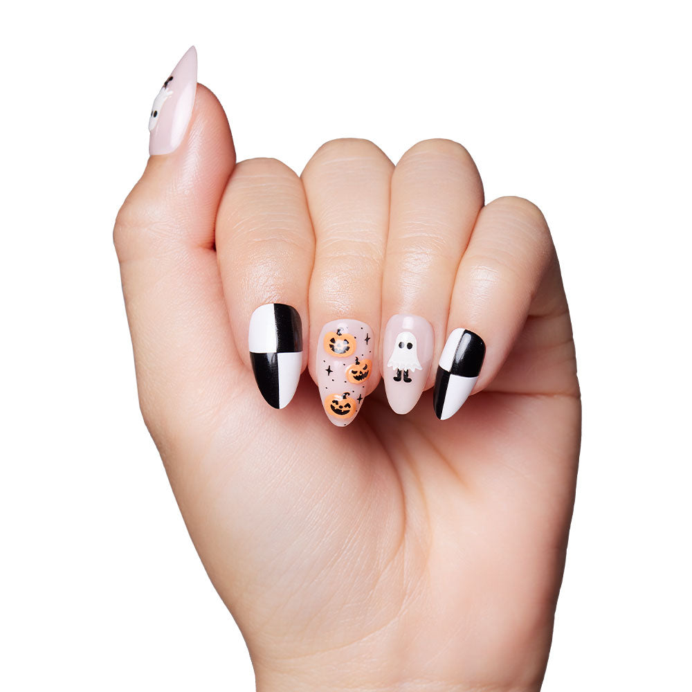 A hand features Halloween-themed press-on nails from glamnetic’s My Boo set, showcasing black and white geometric accents, orange pumpkin faces, and a white ghost design on nude and white nails. The background is white. - Product image