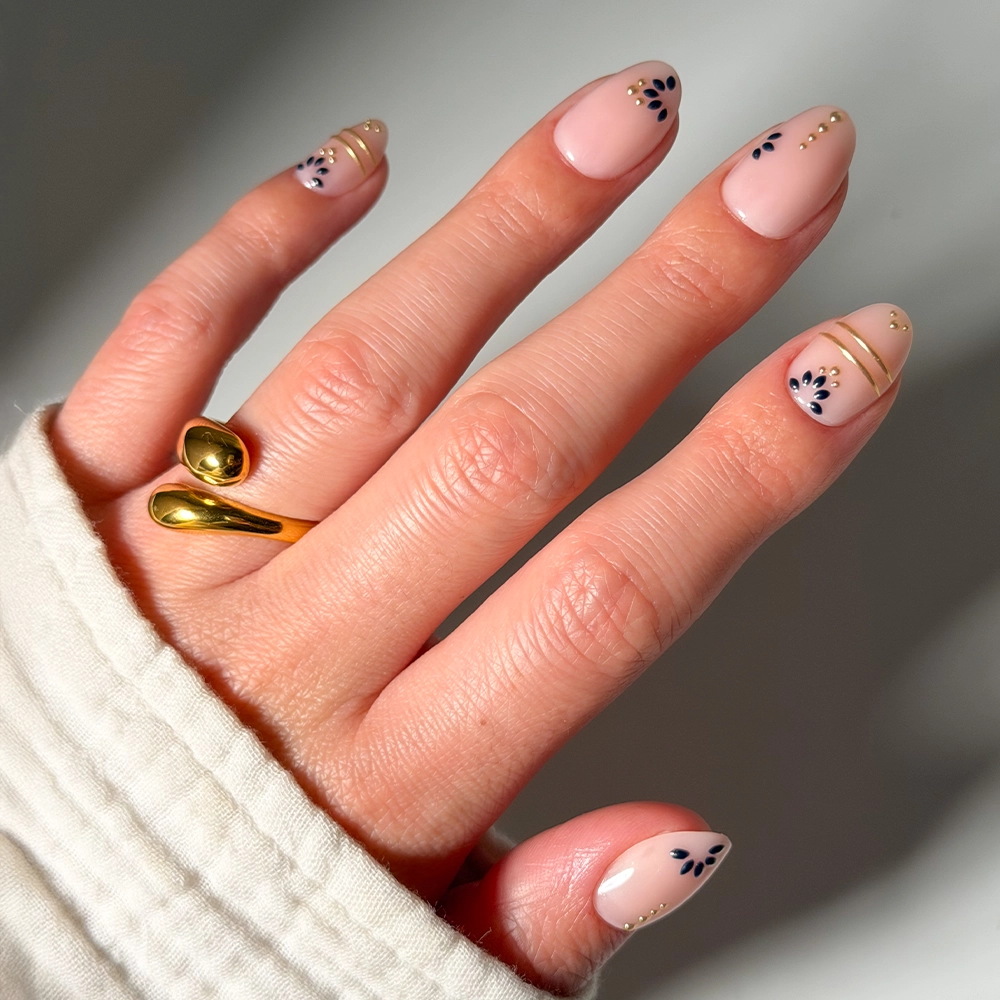 A hand adorned with glamnetic's Myko Muse press-on nails—short ovals with nude polish, black floral art, and gold accents—wears a modern gold ring while a white sleeve partially covers the wrist.