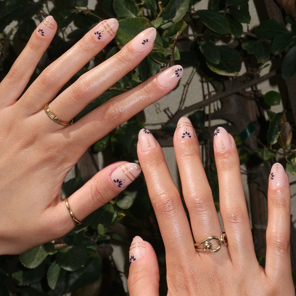 Two hands with short oval nails in Myko Muse by glamnetic show minimalist art—black dots, gold lines, and dainty florals. Gold rings adorn the fingers, set against lush green leafy plants in the background. - Product image