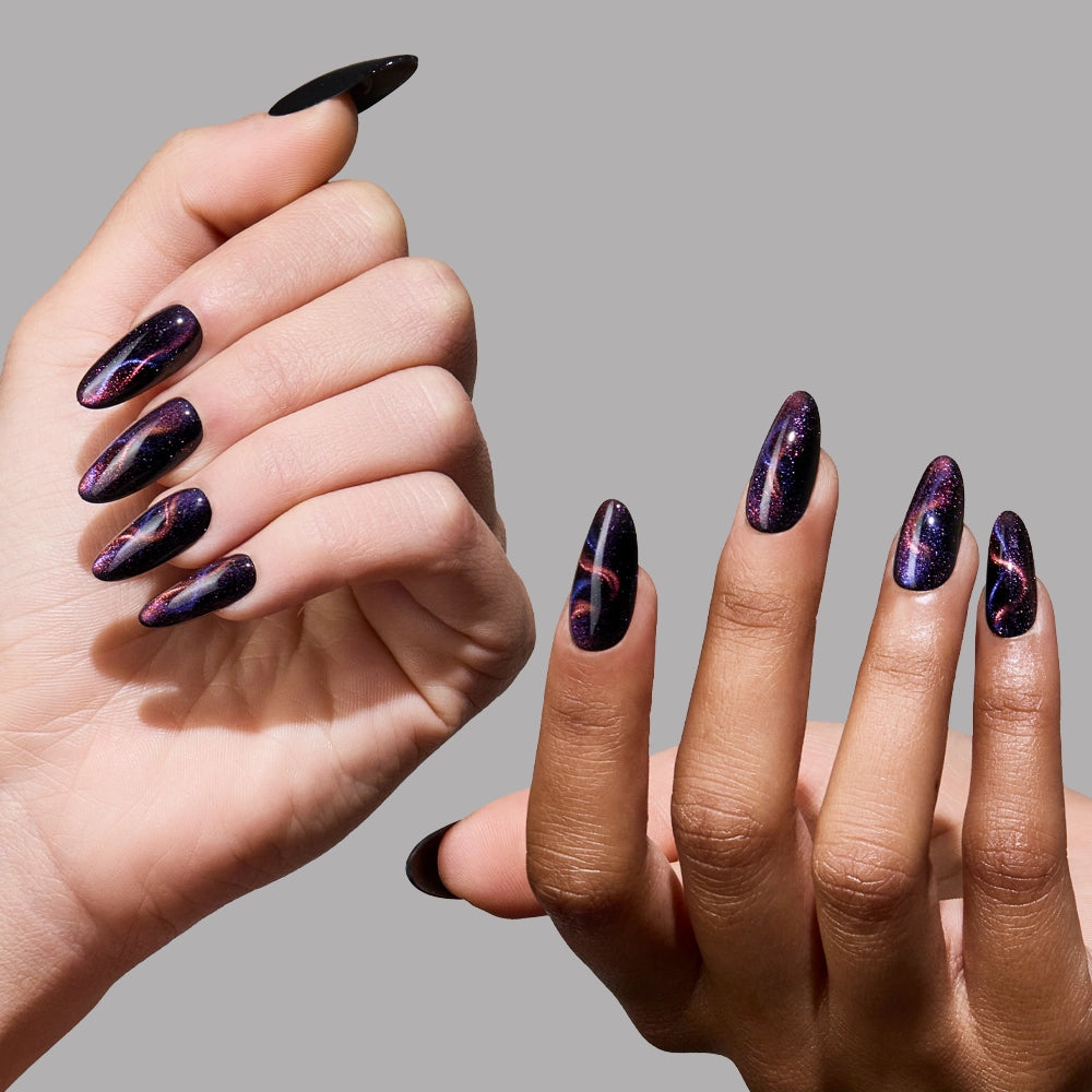 Two hands with almond-shaped nails display glamnetic Mystic Topaz press-ons, featuring a glossy, dark purple and black galaxy design with swirling patterns and sparkles, set against a plain gray background. - Product image