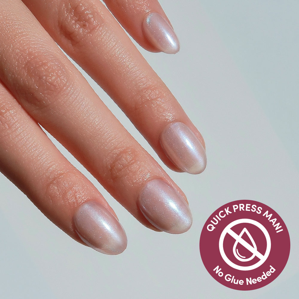 A hand with four fingers visible, featuring glamnetic’s Nude Glazed iridescent stick-on nails. A circular badge reads QUICK PRESS MANI, No Glue Needed.