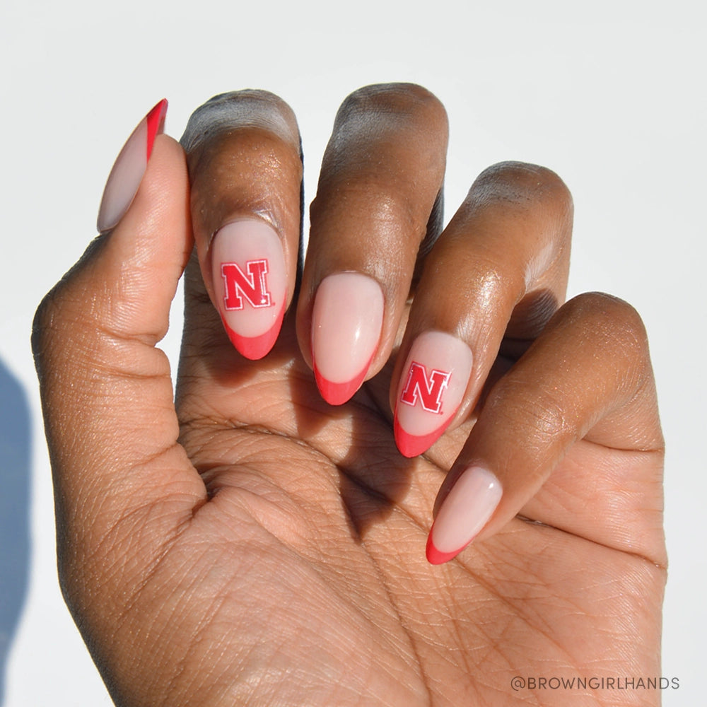 A hand with almond-shaped nails shows the glamnetic Nebraska Cornhuskers Press-On Nail Set: nude base, red French tips, and a red letter N on middle and ring fingers. The background is white.