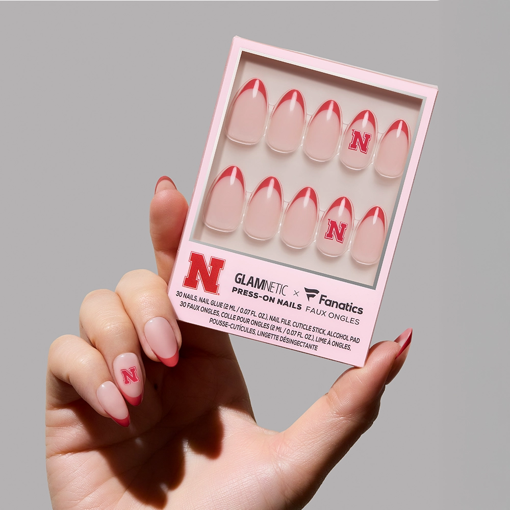 A hand shows off the glamnetic Nebraska Cornhuskers Press-On Nail Set, featuring pink-tipped nails with a red N, displayed in a light pink box. One nail on the hand highlights the signature red N logo.