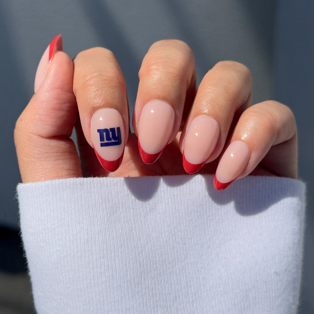 A hand wears glamnetics New York Giants Press-On Nail Set: nude, almond-shaped nails with red French tips and a blue NY logo accent, ideal for NFL nail art. The look is partially covered by a white sleeve.