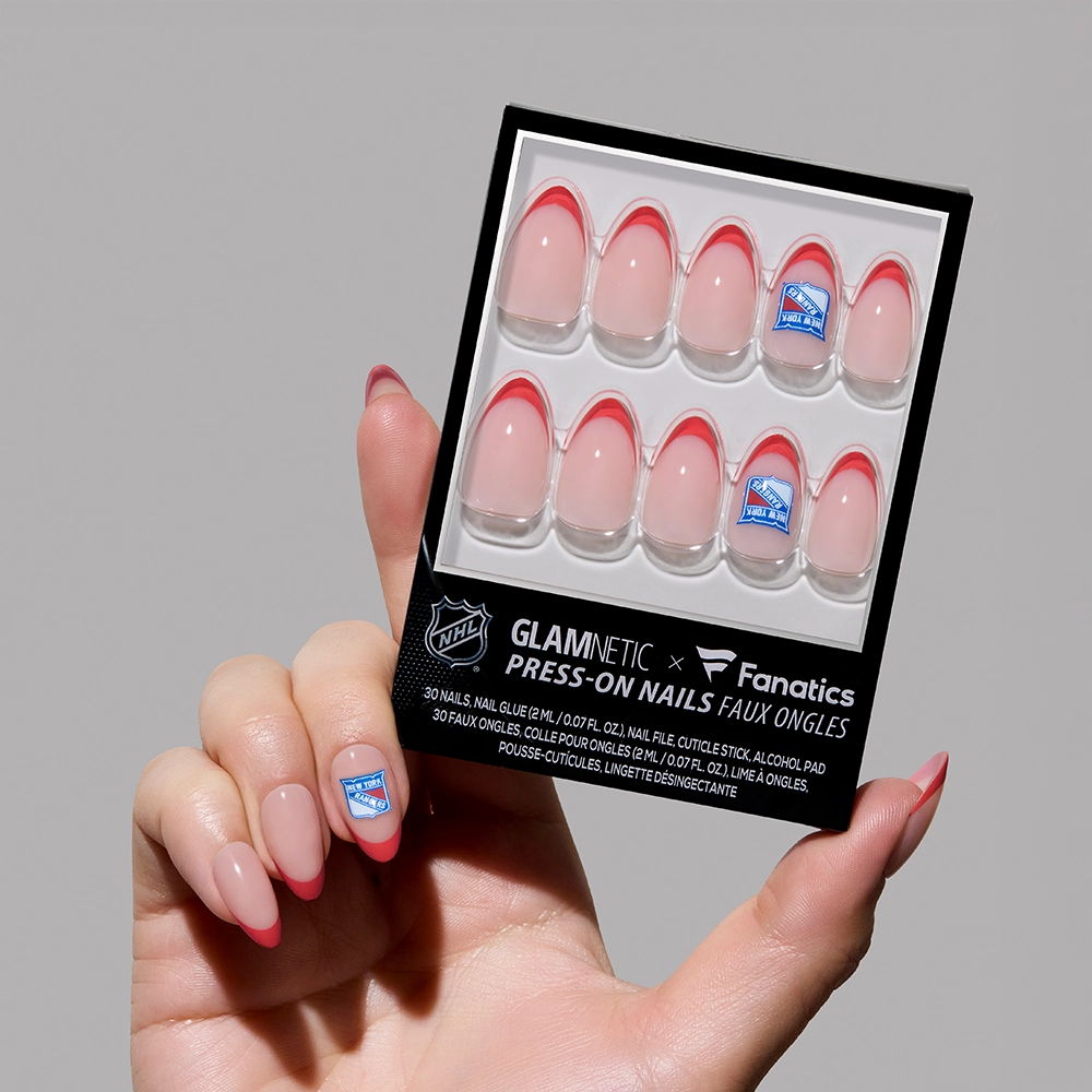 A hand displays the glamnetic New York Rangers Press-On Nail Set with pink French tips. One nail on the hand and two in the pack feature a blue and white Rangers hockey logo. The background is light gray.
