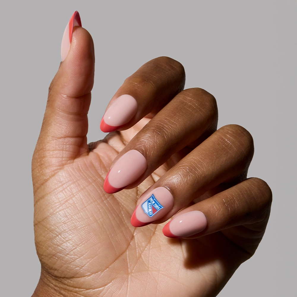 A hand with long, almond-shaped nude nails featuring pink tips and a blue-and-white accent sticker shows off the glamnetic New York Rangers Press-On Nail Set against a plain gray background—ideal for fandom French tip lovers. - Product image