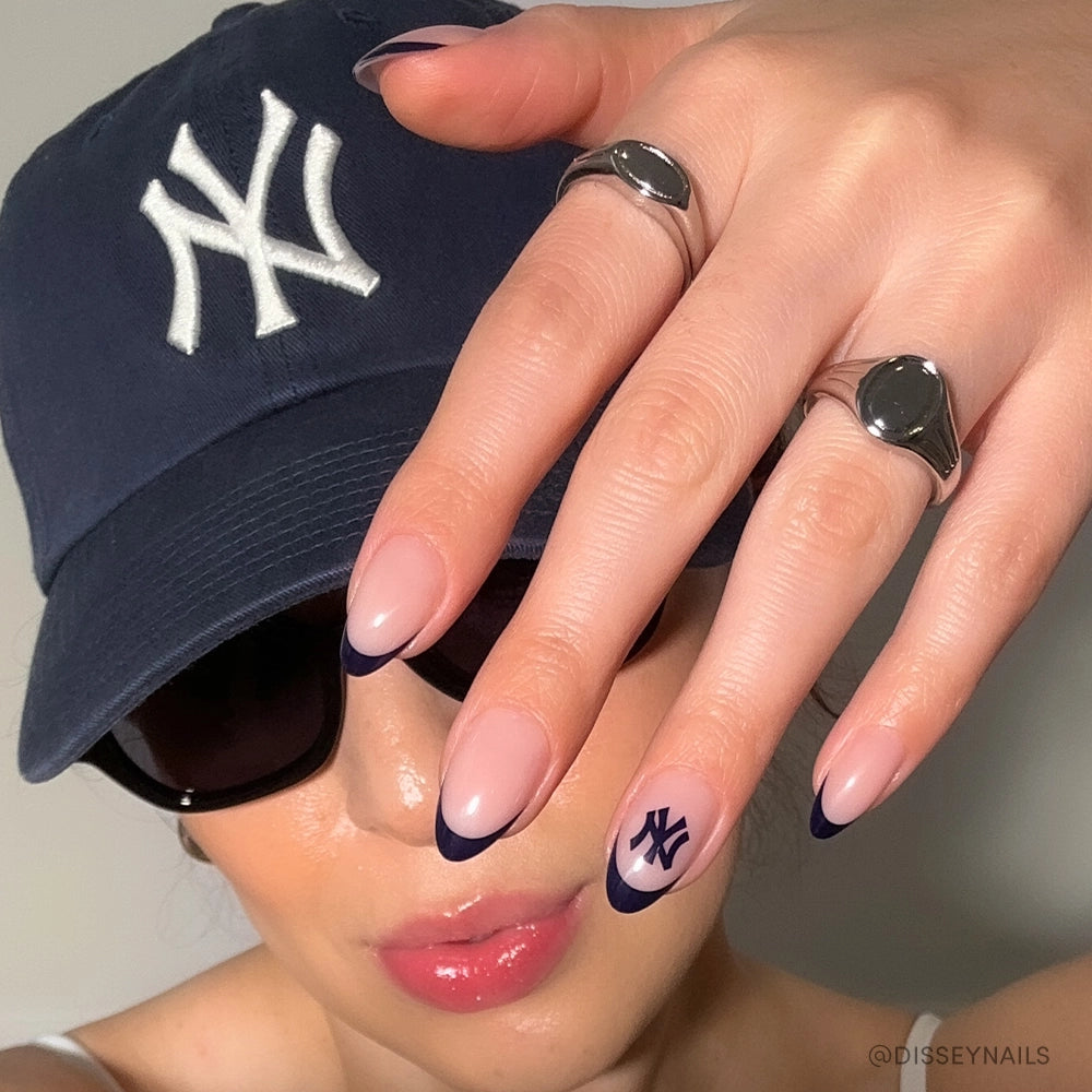 Wearing a navy blue Yankees cap and sunglasses, a person flaunts salon-quality nails with navy blue tips and Yankees logo, featuring the glamnetic New York Yankees Press-On Nail Set.