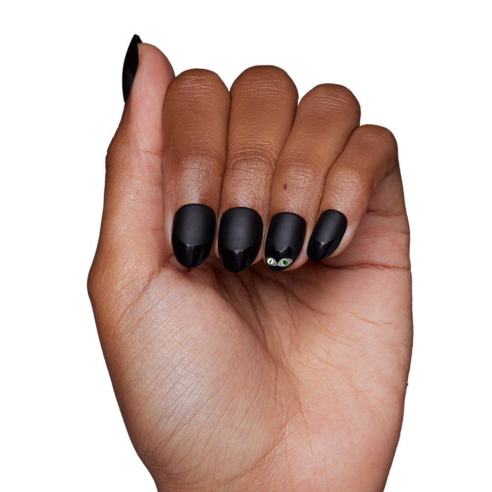 A hand with dark skin displays glamnetics Nine Lives nails in matte black, with two small green gems on the ring finger. The look is striking and cat-inspired, shot against a plain white background. - Product image