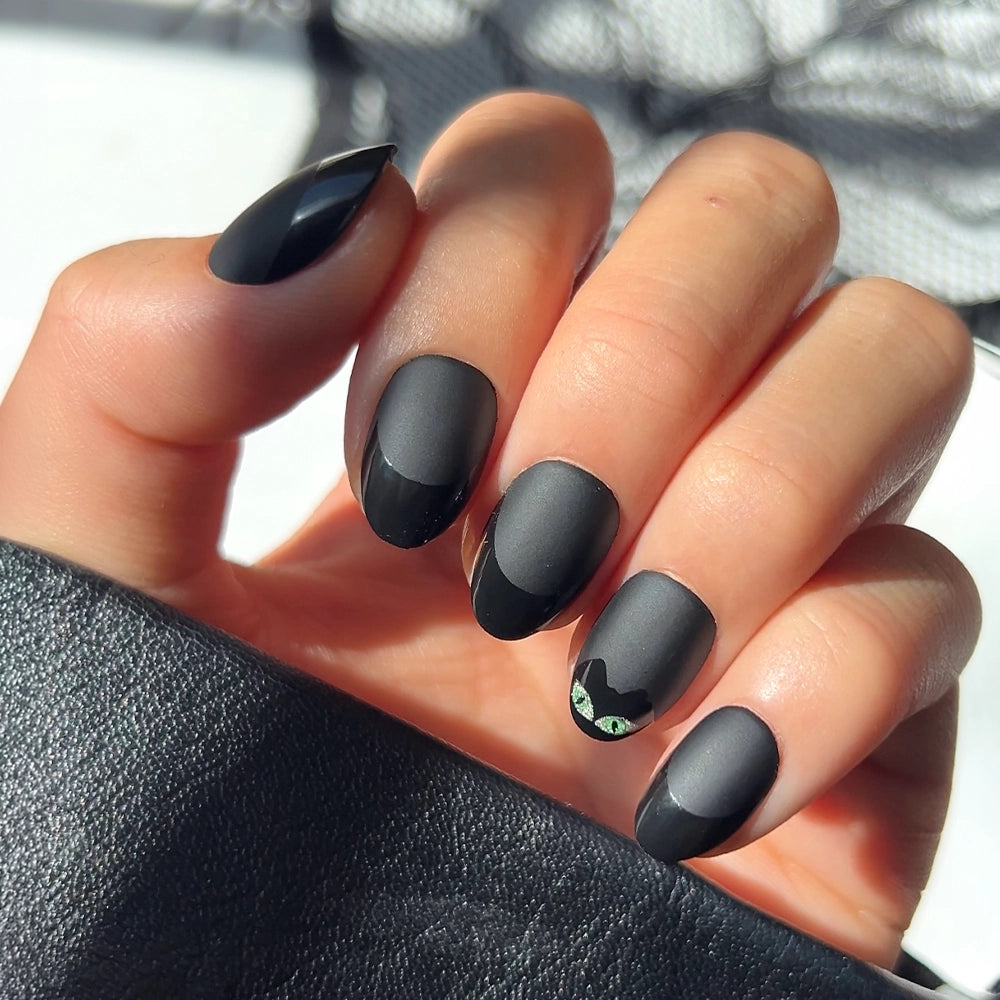 A hand displays the “Nine Lives” press-on nails by glamnetic—short, oval, matte black nails with a striking accent on the ring finger: vivid green cat eyes and black cat ears. The chic black cat design stands out against a sleek black sleeve.