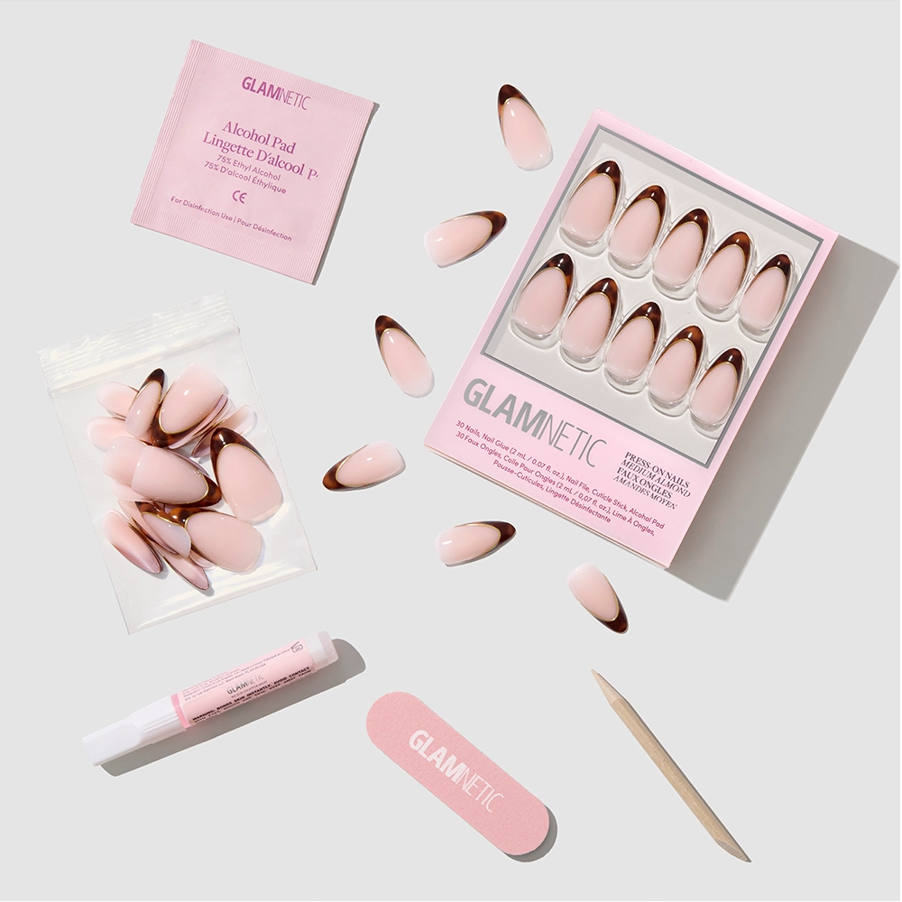 The "No Competition" almond-shaped pink and gold press-on nails by glamnetic come with nail glue, a wooden cuticle stick, mini file, alcohol pad, and extra nails—all arranged neatly on a white background. - Product image