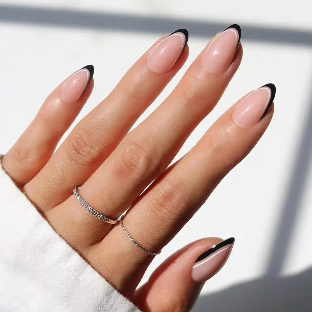 A hand with long almond-shaped nails painted in glamnetic’s NO IN BETWEEN—a nude shade with micro black French tips—displays two delicate silver rings and a white long-sleeve top, set against a bright, softly blurred background.