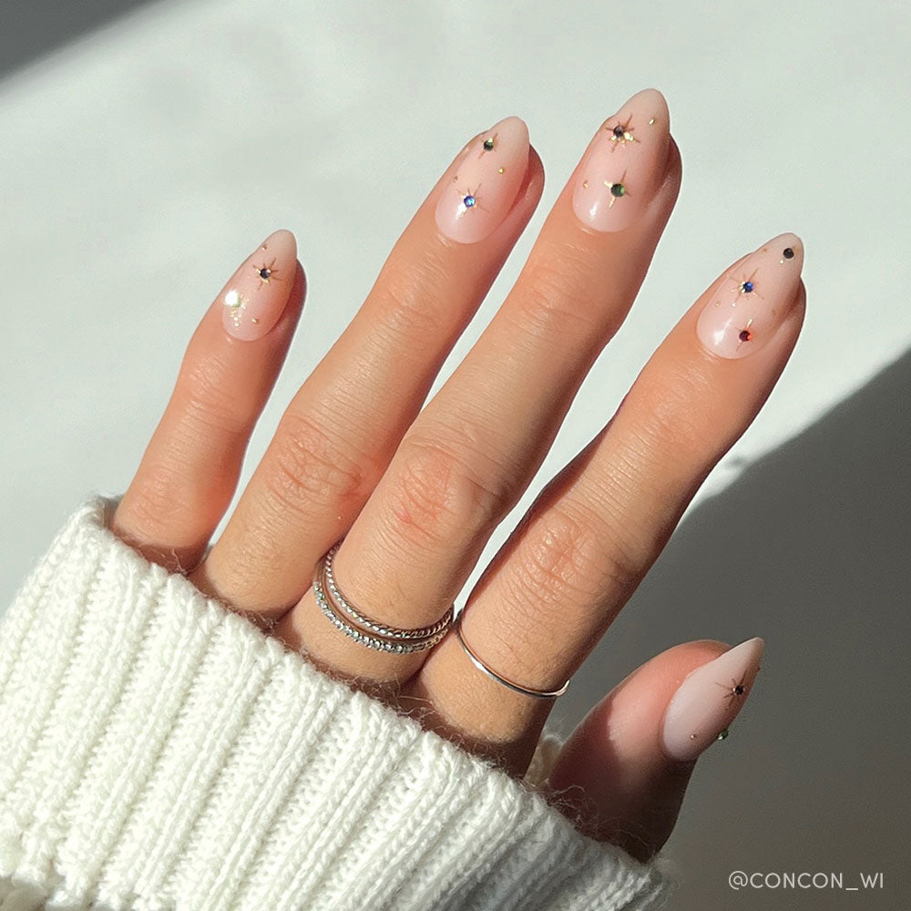 A hand with long, almond-shaped nails featuring glamnetic’s North Star design—nude polish with colorful star accents and gold foil details. The look is complemented by silver rings and a white knit sweater.