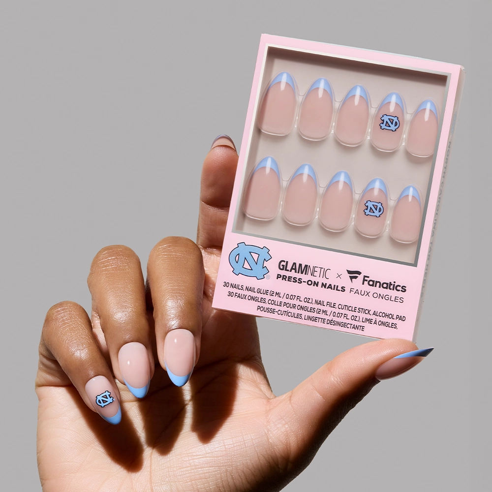A hand with North Carolina Tar Heels Press-On Nail Set by glamnetic, featuring Carolina-blue French tips and the UNC logo, holds a glamnetic box showing more salon-quality, UNC-themed press-on nails.