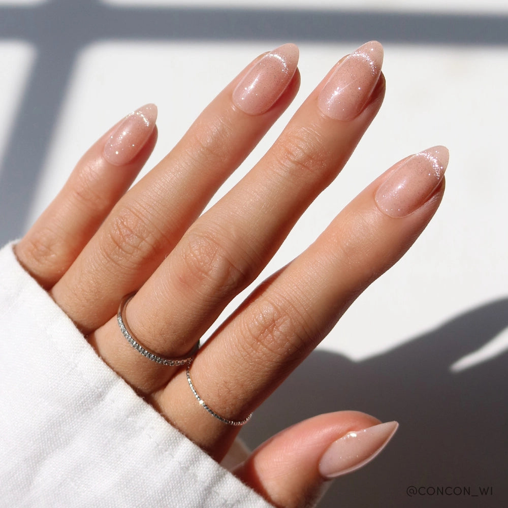 Hand wearing almond-shaped nude shimmer press-on nails with subtle glitter and French tip effect, accessorized with silver rings.