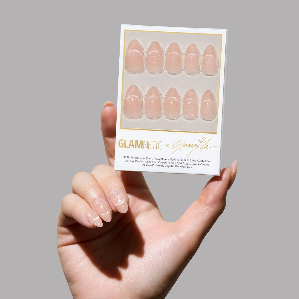Hand holding Glamnetic x Glamzilla “Not Your Cinder-Ella” press-on nail set featuring 30 nude shimmer nails in various sizes.