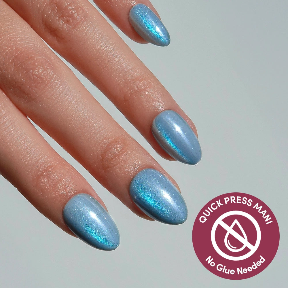 A hand with almond-shaped, metallic blue glamnetic Ocean Prism press-on nails. A badge reads QUICK PRESS MANI No Glue Needed with a crossed-out glue bottle, showcasing these easy-to-apply stick-on nails.