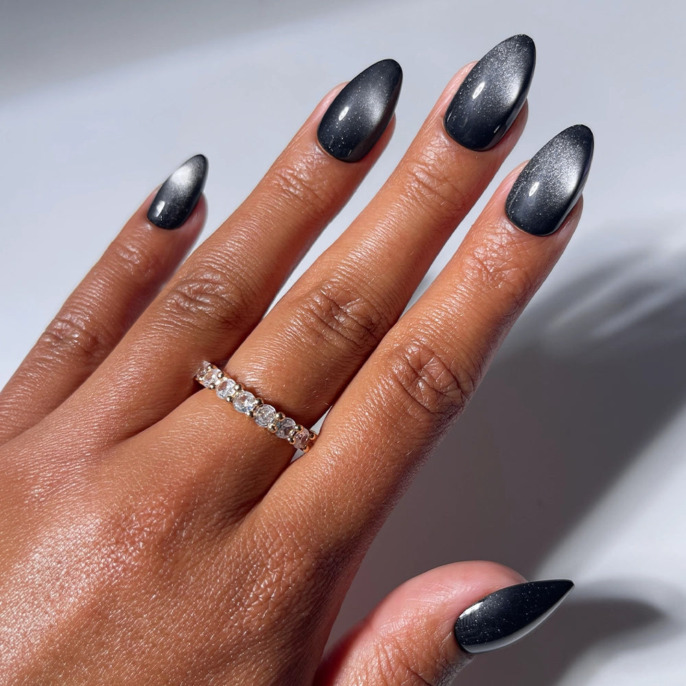 A hand with long, almond-shaped glamnetic Obsidian Press-On Nails and a sparkling diamond ring on the ring finger, set against a plain light background.