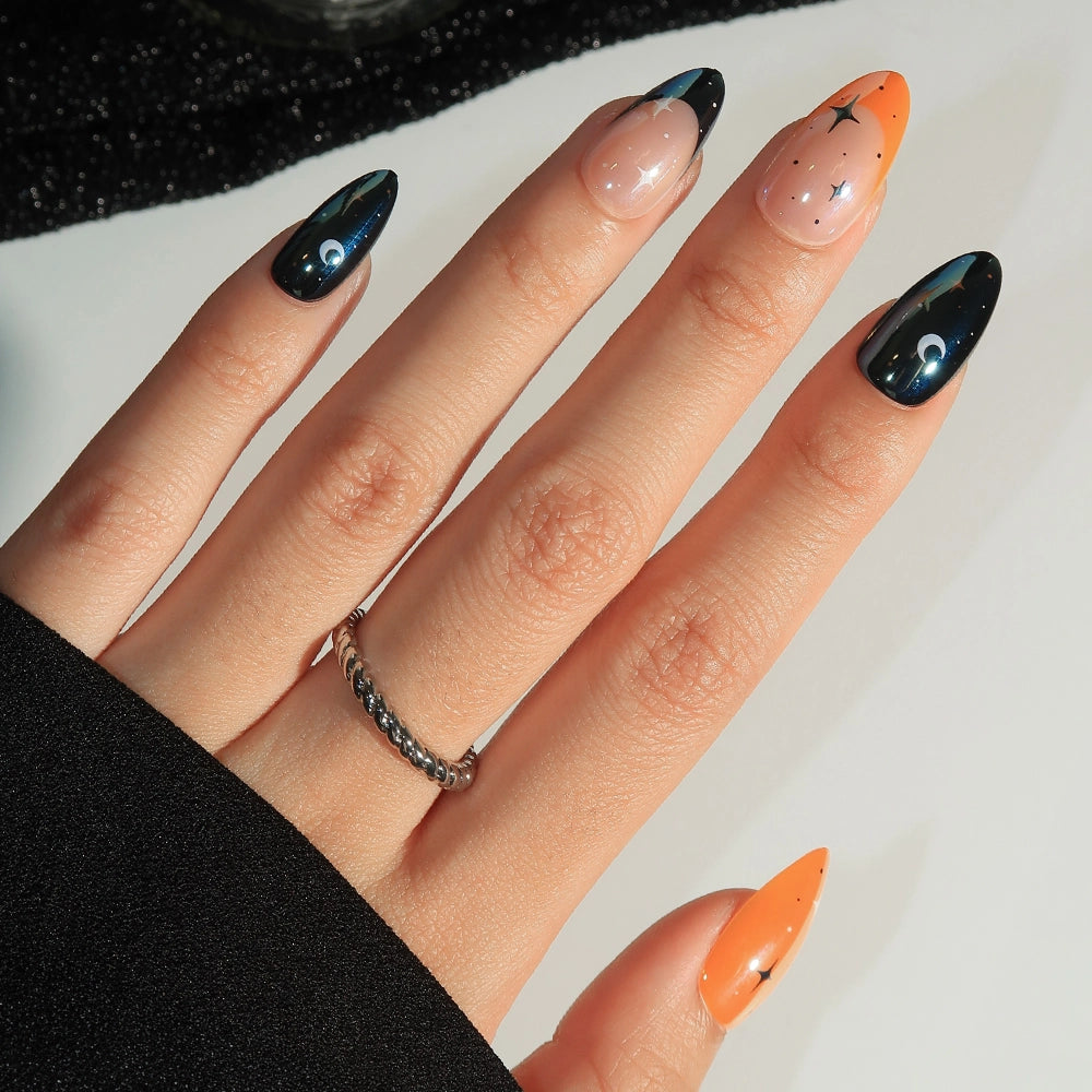 A hand showcases “October Nights” press-on nails by glamnetic, featuring long, almond-shaped tips painted in a cosmic theme with stars, moons, and gradient shades of black, peach, and orange. Some nails are accented with glazed French tips for added shine. The look is completed with a silver twisted ring on the middle finger. - Product image