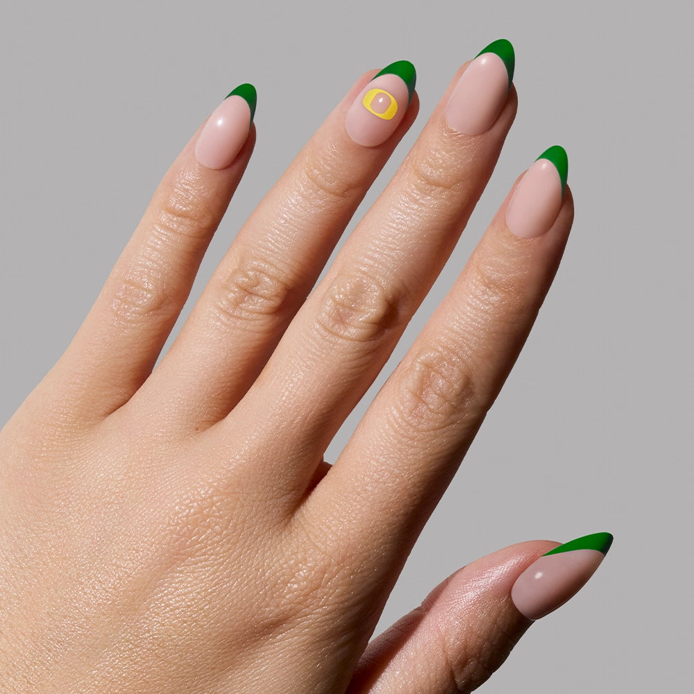 A hand with almond-shaped green French tip nails from the glamnetic Oregon Ducks Press-On Nail Set; the ring finger has a yellow square with a small white circle. Shown against a plain light gray background. - Product image