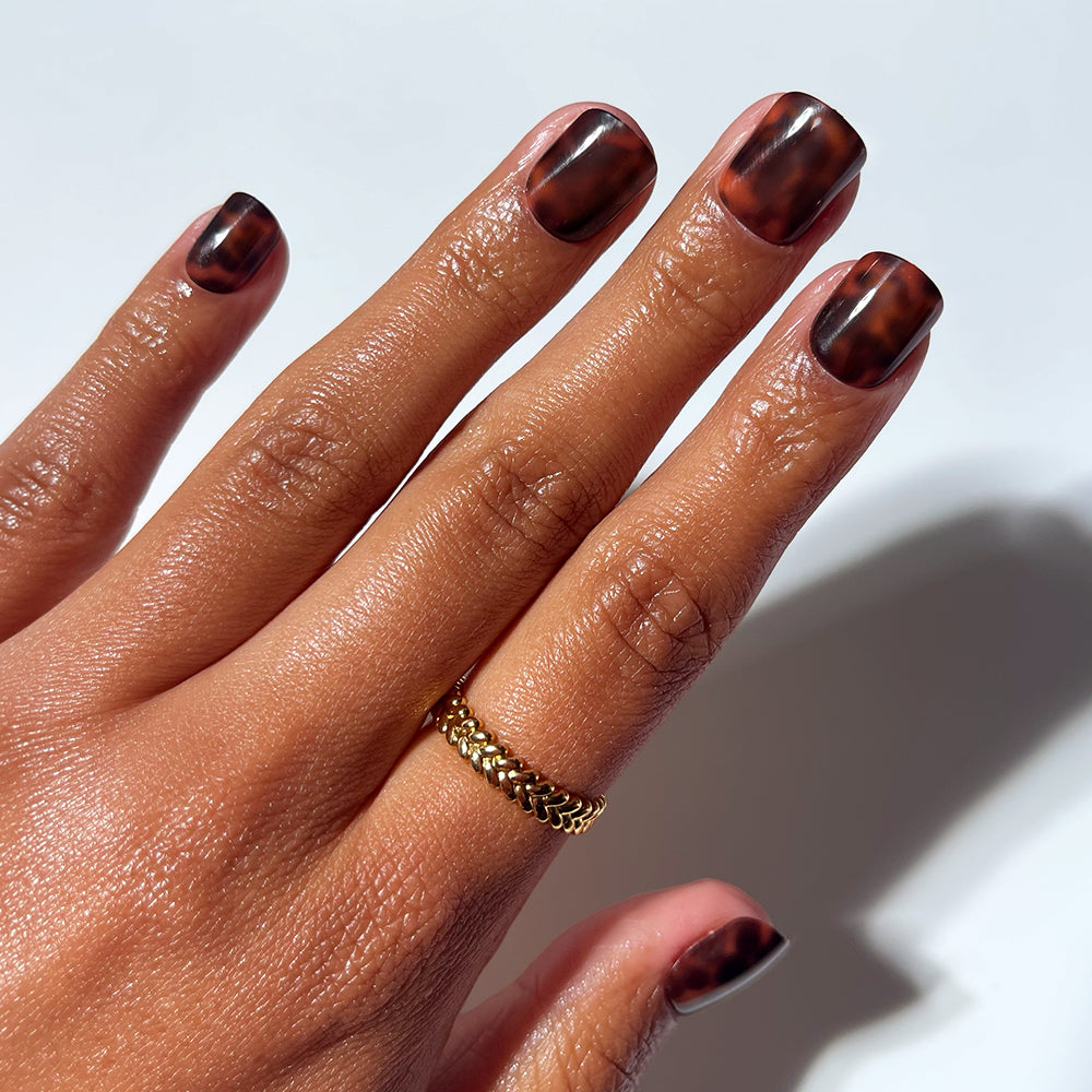A hand with short, square glamnetic "Own It All" press-on nails in a glossy brown tortoiseshell design, paired with a gold chain-link ring on the middle finger, set against a white background.