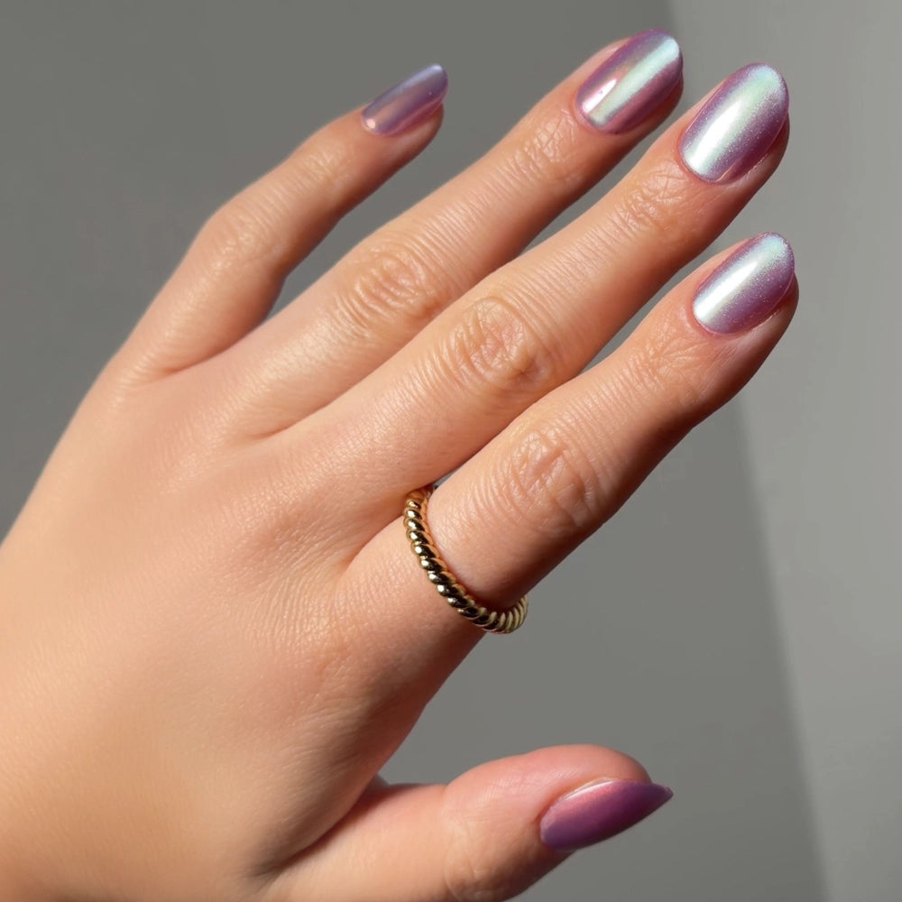 A hand displays glamnetic's Oyster Glow iridescent light purple almond-shaped press-on nails. A gold beaded ring accents the ring finger, while a simple gray backdrop highlights the pearlescent nails and jewelry.