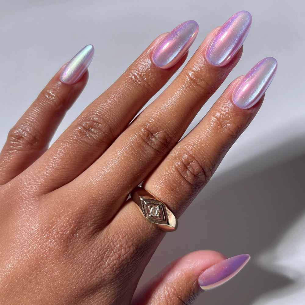 A hand with medium oval nails painted in glamnetic's Oyster Pearl, a shimmering iridescent pink. A gold ring with a small diamond adorns the ring finger, set against a soft white background.