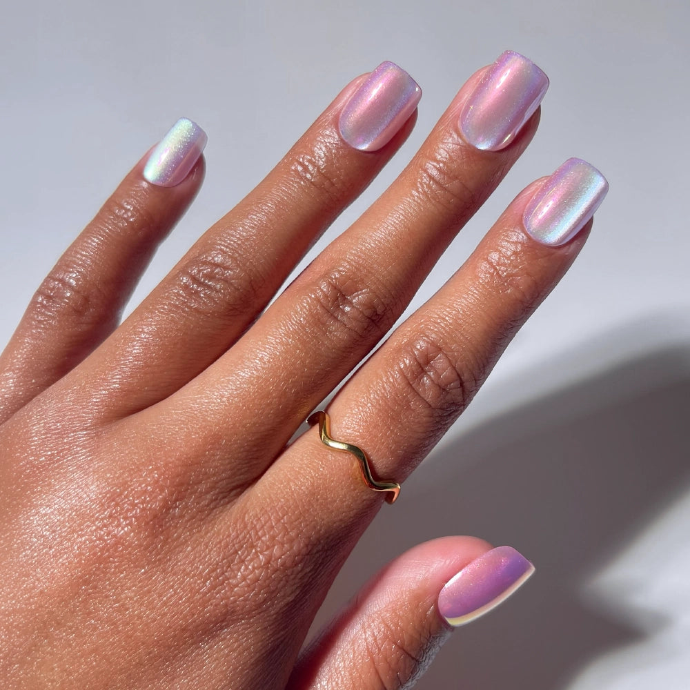 A hand with glamnetic’s Oyster Shell iridescent pastel pink and purple short squoval nails wears a simple gold wavy ring on the middle finger, set against a plain light background.