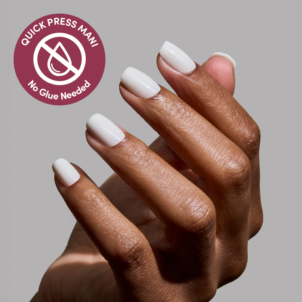 A hand with medium-brown skin showcases glossy white press-on nails in "Porcelain" by glamnetic. A circular label says Quick Press Mani, No Glue Needed, with a crossed-out droplet. The light gray background highlights these easy, no-glue nails. - Product image
