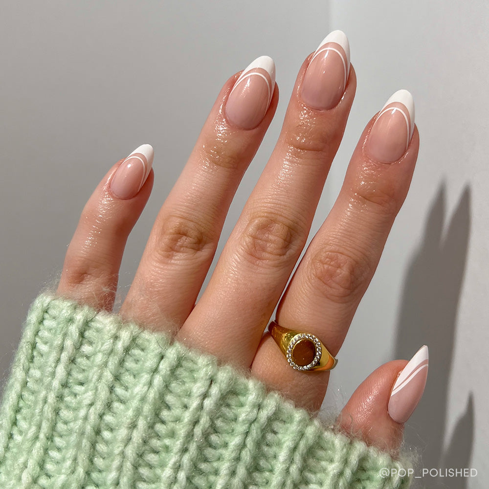 A short almond hand flaunts glamnetic’s Paris nails—a modern white French tip with fine white lines. The look radiates French sophistication, styled with a gold ring (brown stone) and a pale green knit sweater. - Product image