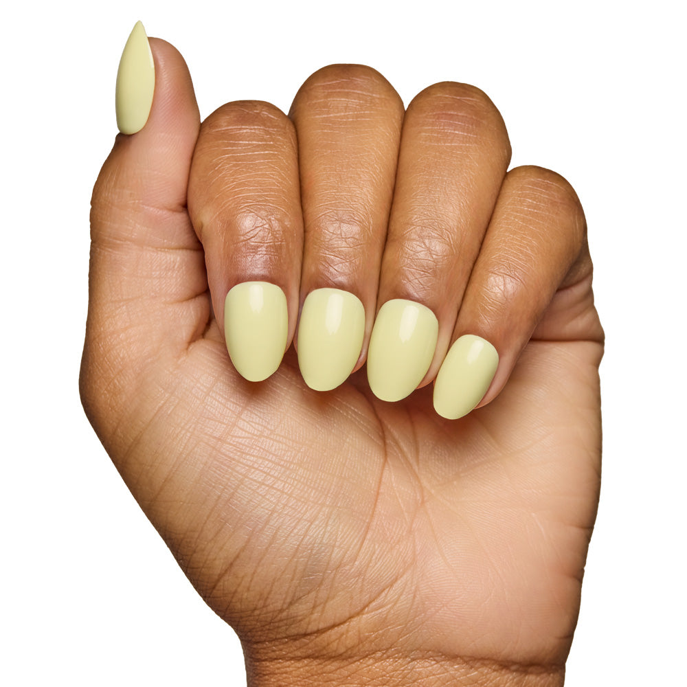 A hand with medium brown skin displays short oval nails in glamnetics Pastel Yellow, featuring a glossy, salon-quality finish. The fingers curl slightly against a white background, emphasizing the rounded almond shape. - Product image