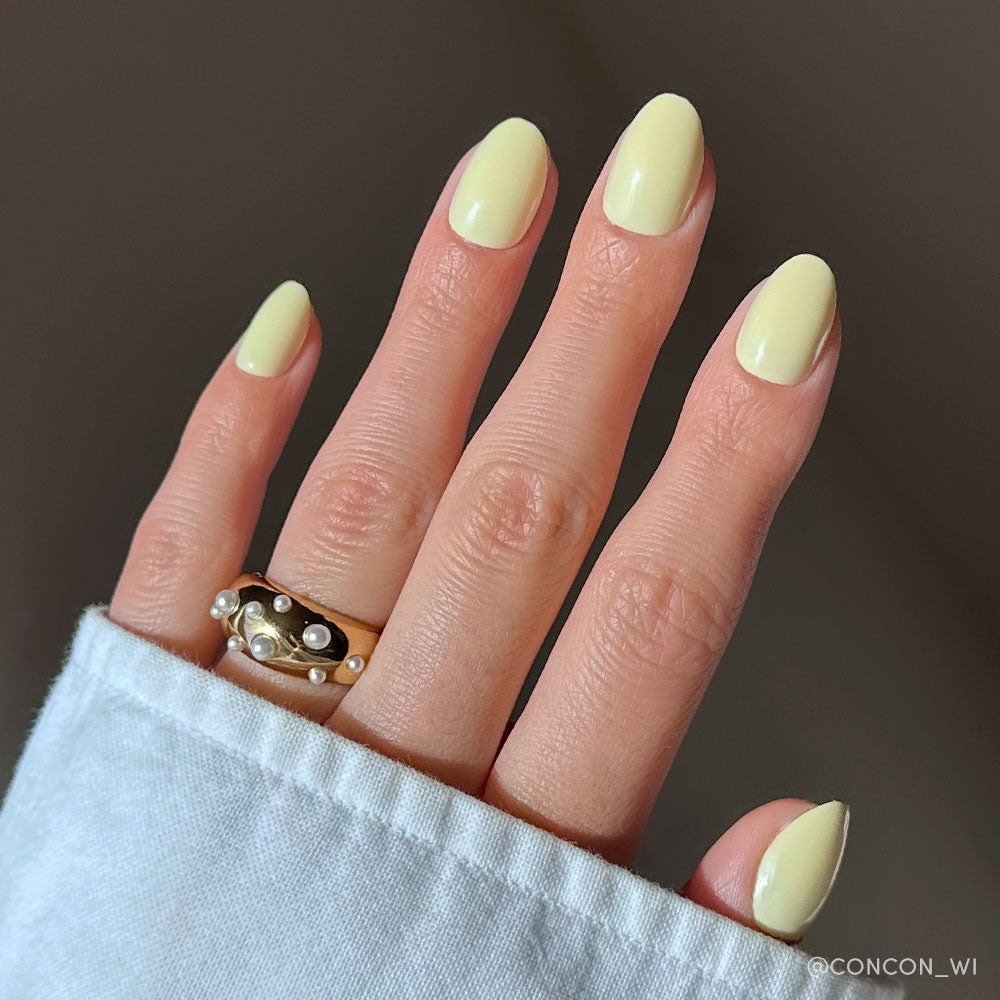 A hand with salon-quality sheen on short oval nails, painted in glamnetics Pastel Yellow set, wears two gold rings with pearl accents. The person is dressed in a light-colored long-sleeve shirt against a neutral background. - Product image