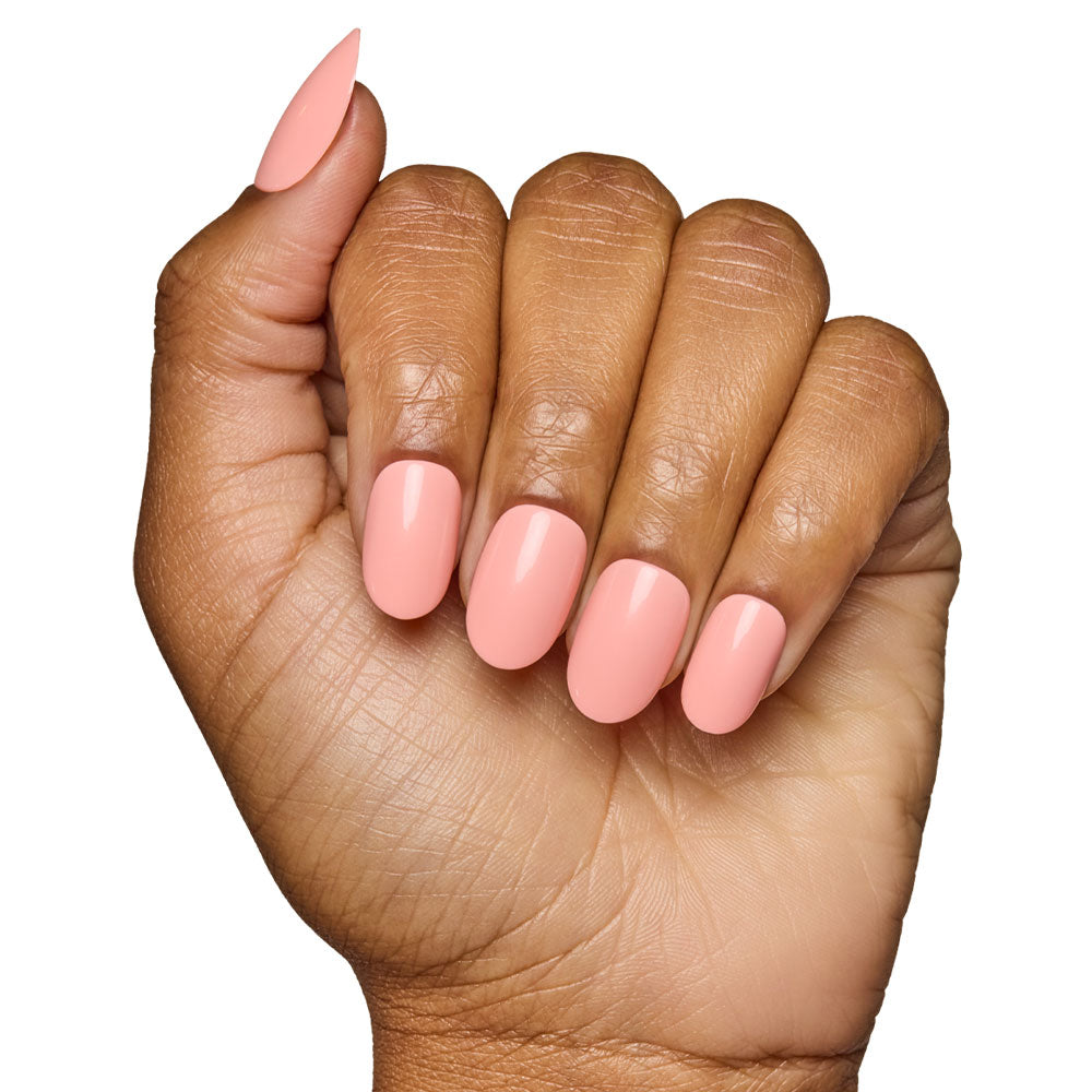 A hand with medium brown skin displays almond-shaped nails coated in glamnetic’s Peach Coral, a glossy pastel peach shade. The pinky nail is longer and more pointed, echoing the elegance of the Peach Coral Nail Set. - Product image
