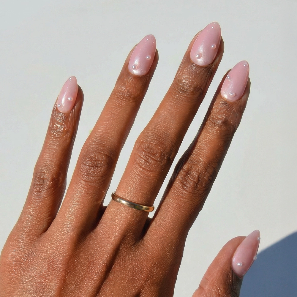 A hand with medium brown skin wears a simple gold ring on the middle finger. Long, blush-pink almond nails are styled with glamnetic's Pearl Me Softly set, featuring delicate 3D pearl accents for an elegant touch.