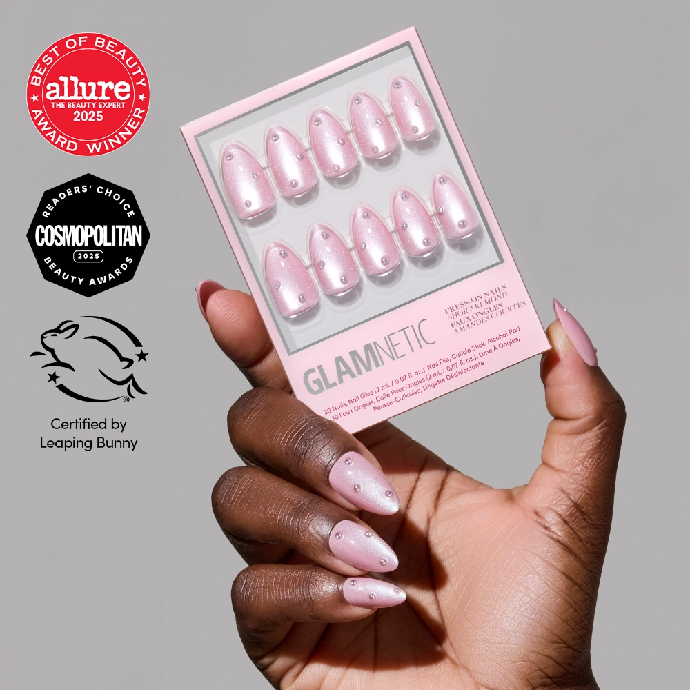 A hand with light brown skin holds a box of glamnetic Pearl Me Softly blush-pink almond nails, also worn on the hand. Beauty award logos from Allure, Cosmopolitan, and Leaping Bunny appear on the left.