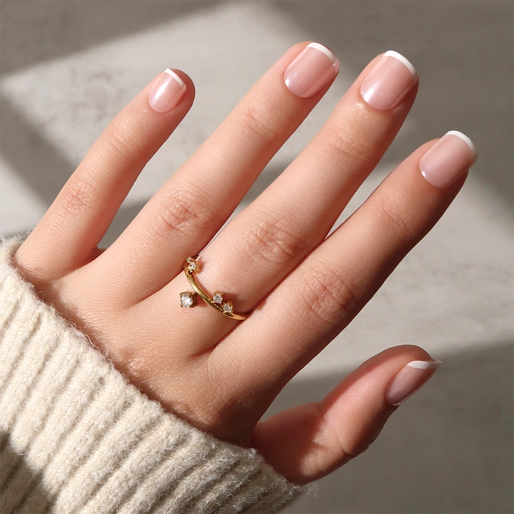 A hand with a light skin tone and a French tip manicure wears the glamnetic "Pearly Please" gold ring adorned with small gemstones. The beige ribbed sweater and softly blurred background enhance the look’s elegance. - Product image