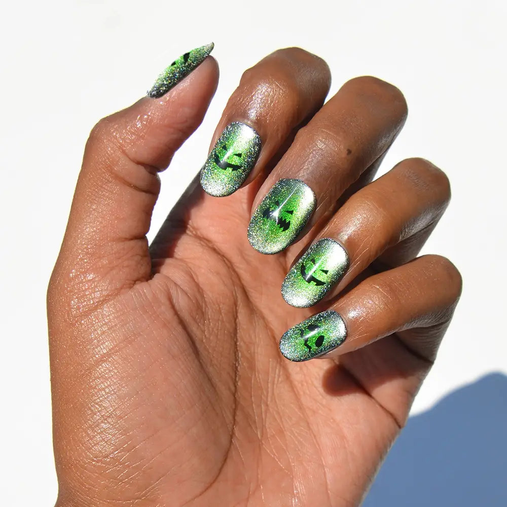 A hand with long, manicured Peek-o’-Lantern nails from glamnetic features a silver and green velvet shimmer finish with cat-eye inspired black designs. The background is white. - Product image