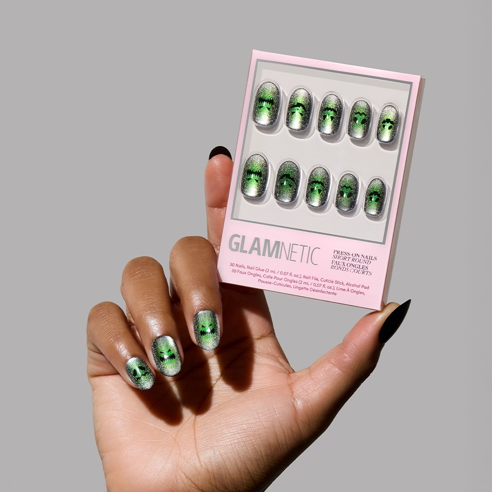 A hand with magnetic green velvet shimmer, short round ombré Peek-o’-Lantern press-on nails holds a matching glamnetic box against a plain gray background, highlighting the reusable manicure.