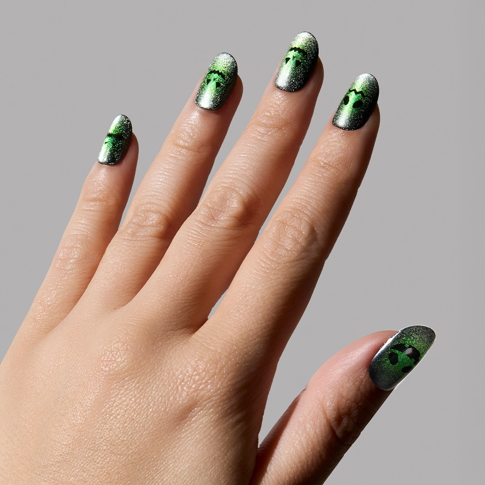 A hand with short round nails wears glamnetic’s Peek-o’-Lantern: black and magnetic green velvet gradient nails with tiny alien faces, shown against a plain gray background. - Product image