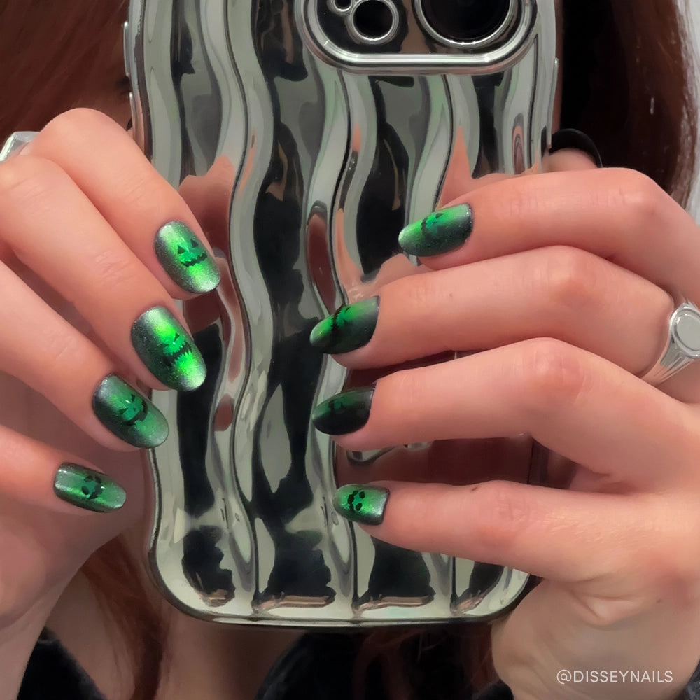 A person wears glamnetic Peek-o’-Lantern reusable manicure on short round nails with green and black holographic jack-o-lantern faces, holding a shiny, wavy silver phone case. Silver rings adorn their fingers. - Product image