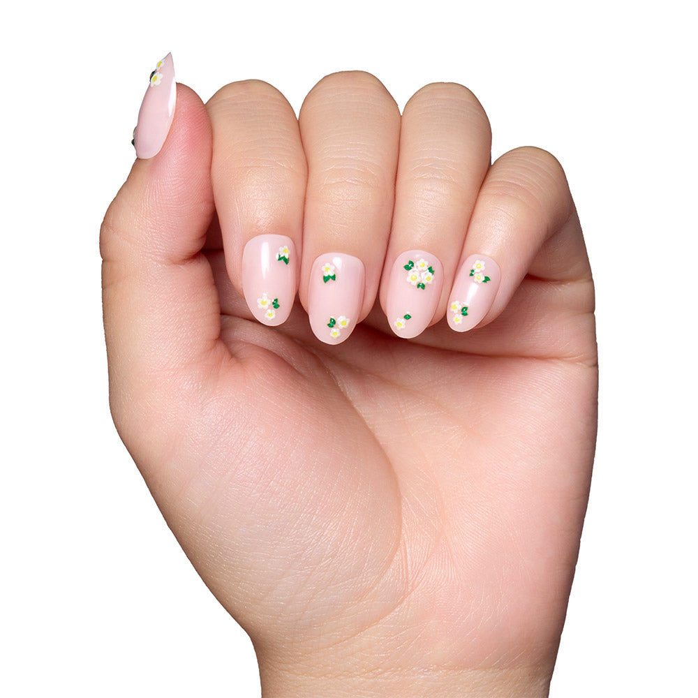 A hand with short oval nails wears glamnetics Petals: pale pink, glossy press-ons with delicate white and yellow floral accents, shown against a plain white background. - Product image