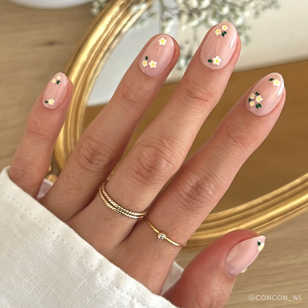 A hand with glamnetics Petals press-on nails—light pink with a glossy finish and white-yellow daisy accents on short ovals—is shown, styled with two gold rings and a white cuffed sleeve.