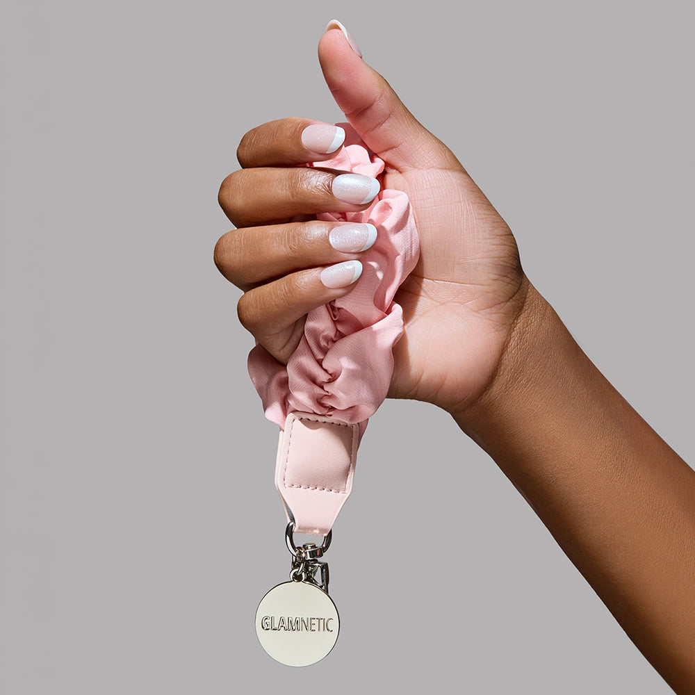 A hand with a light pink manicure holds a pink Glamnetic Phone Wristlet keychain by glamnetic, featuring a round silver tag, against a plain gray background. - Product image