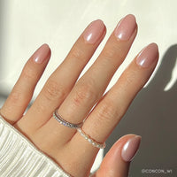 A hand with neatly manicured, short oval nails in glamnetics Pinch Me light pink shade with a glossy glaze, adorned with a silver diamond ring and pearl ring, wearing a white ribbed sweater.