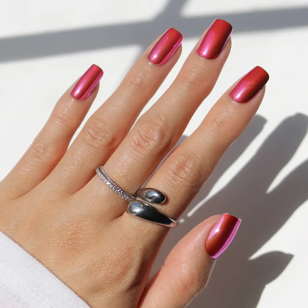 A hand showcases glamnetic’s Pink Alloy—medium square press-on nails in a shiny, metallic red-pink chrome finish—adorned with two silver rings: one with a wrap design and the other embellished with small crystals, all set against a white background. - Product image