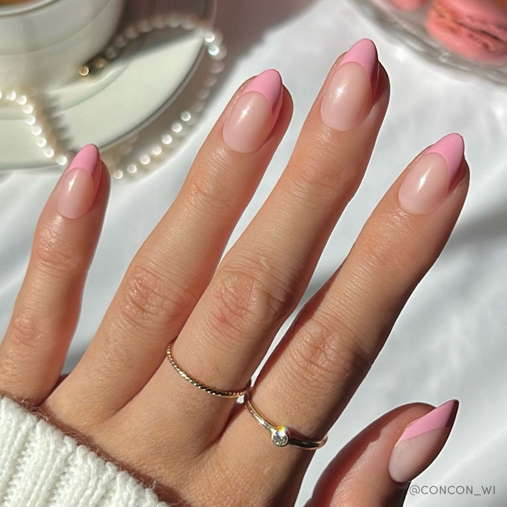 A hand with an almond base wears glamnetics Pink Macaron nails in pink French tips. Two delicate gold rings adorn the fingers, and a cup with a pearl necklace appears in the background. - Product image